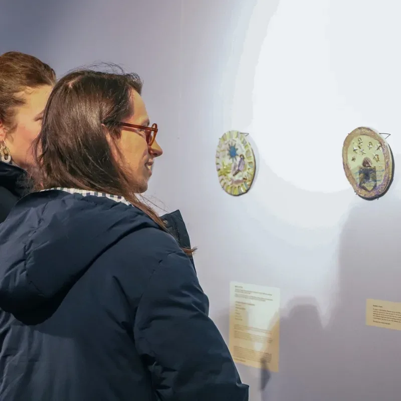 Two women in an exhibition looking at ceramic plaques on a white wall.