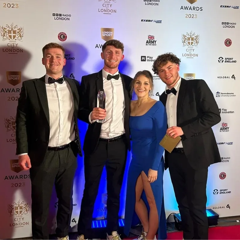 Four people pose together at an awards ceremony. Three men are wearing tailored suits, while a woman stands beside them in an elegant dress. They all appear happy and are smiling for the camera.