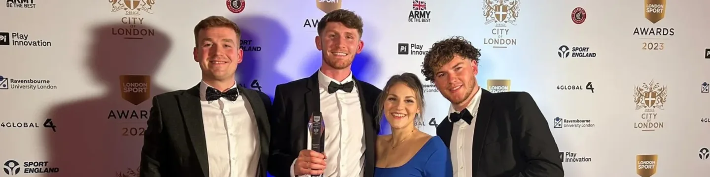 Four people pose together at an awards ceremony. Three men are wearing tailored suits, while a woman stands beside them in an elegant dress. They all appear happy and are smiling for the camera.