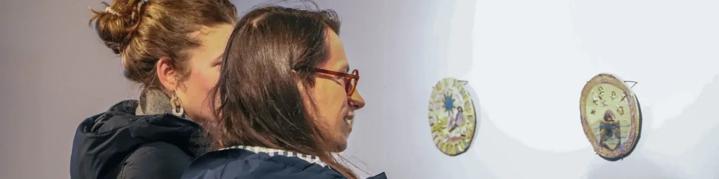 Two women in an exhibition looking at ceramic plaques on a white wall.