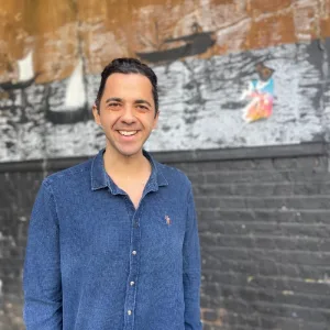 A man in a blue shirt stands on the street, grinning in front of a black brick wall.