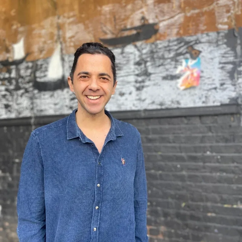 A man in a blue shirt stands on the street, grinning in front of a black brick wall.