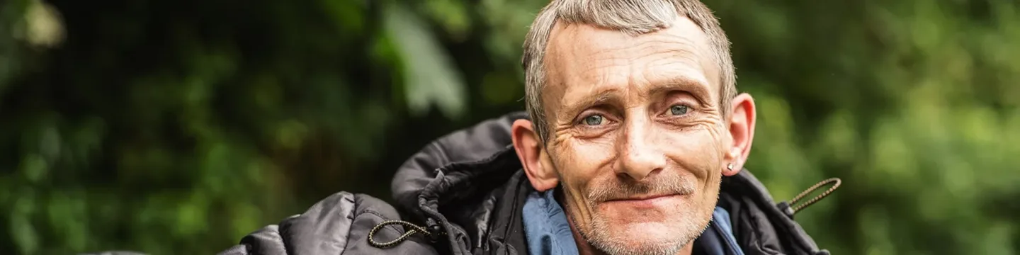 An older man sits on a park bench, wearing a black puffer jacket, with greenery in the background.