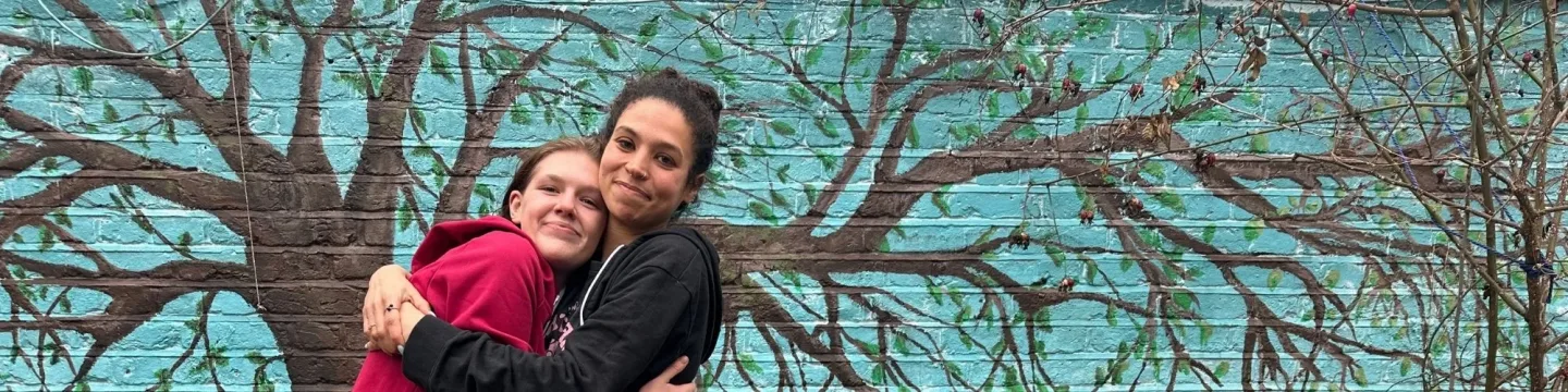 Two women hug in front mural of tree, showing mutual respect and love