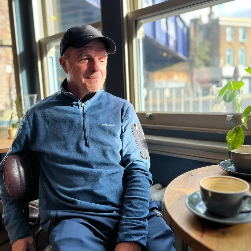 An older man sits in a leather chair at a coffee shop with two cups and a plant on the table in front of him. He is dressed in a blue jumper and trousers and wears a black hat.