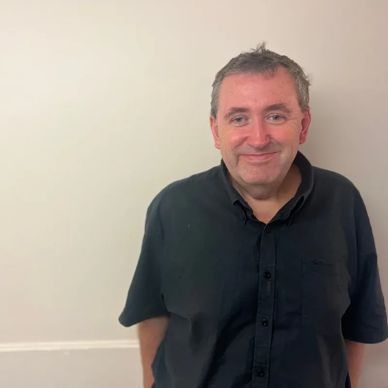 An older man with a cheerful grin stands in front of a cream-coloured wall, wearing a black shirt.