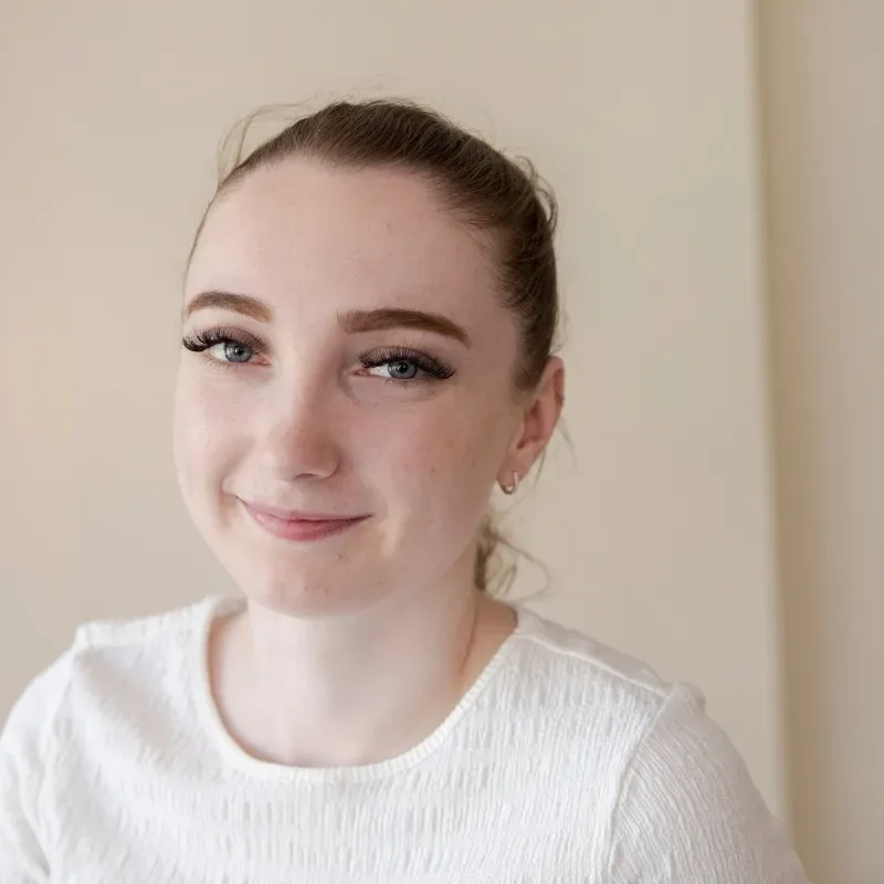 A young woman in a white top smiles while standing indoors, with a cream wall in the background.