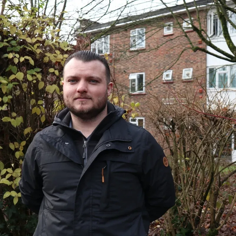 A man wearing a black jacket with slicked-back hair stands with trees and houses in the background.