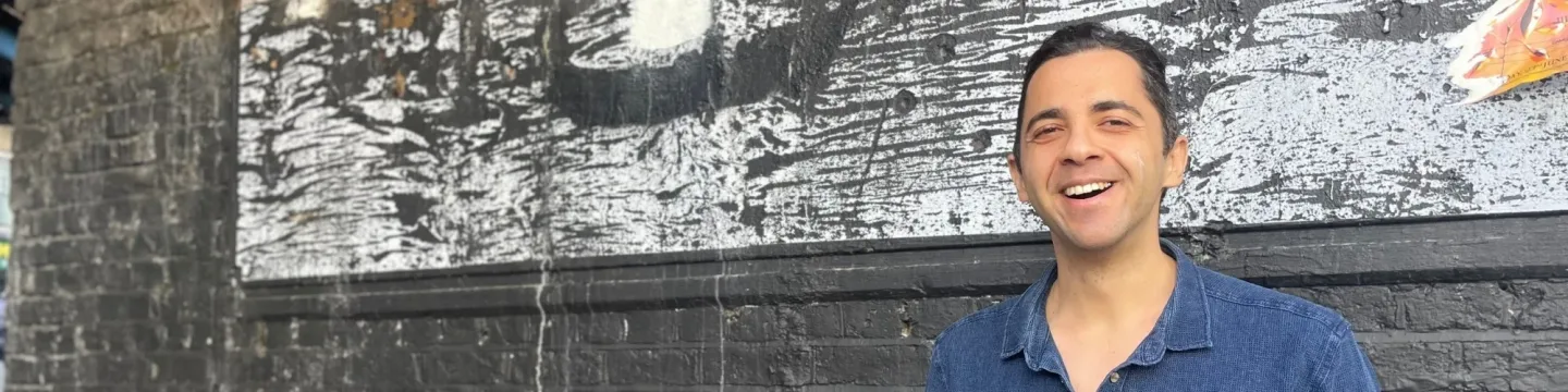A man in a blue shirt stands on the street, grinning in front of a black brick wall.