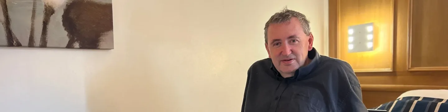 An older man sits on a bed in a hotel room, wearing a black shirt, with a cream-coloured wall and a painting visible behind him.