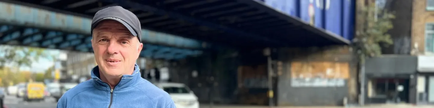 An older man stands on the street with Camden Road bridge visible behind him. He is wearing a blue jumper and a black hat.