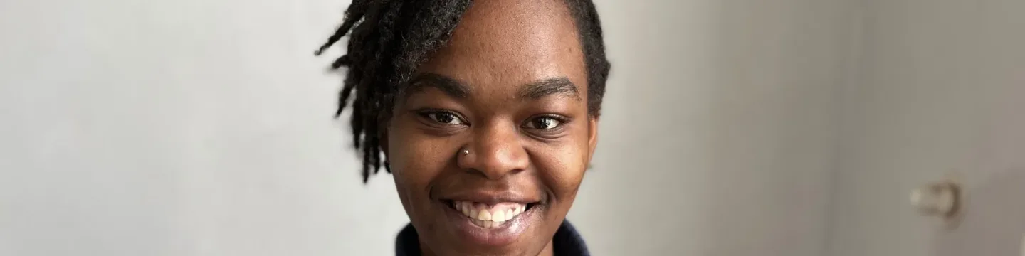 A woman in a red jumper stands indoors in front of a cream-colored wall.