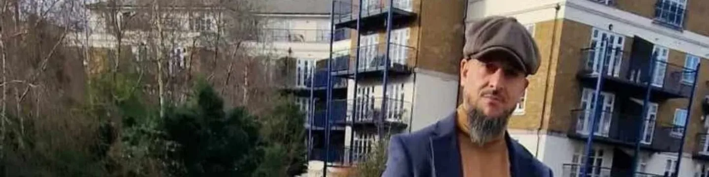 A man wearing a hat and a trench coat stands on a bridge overlooking a canal.