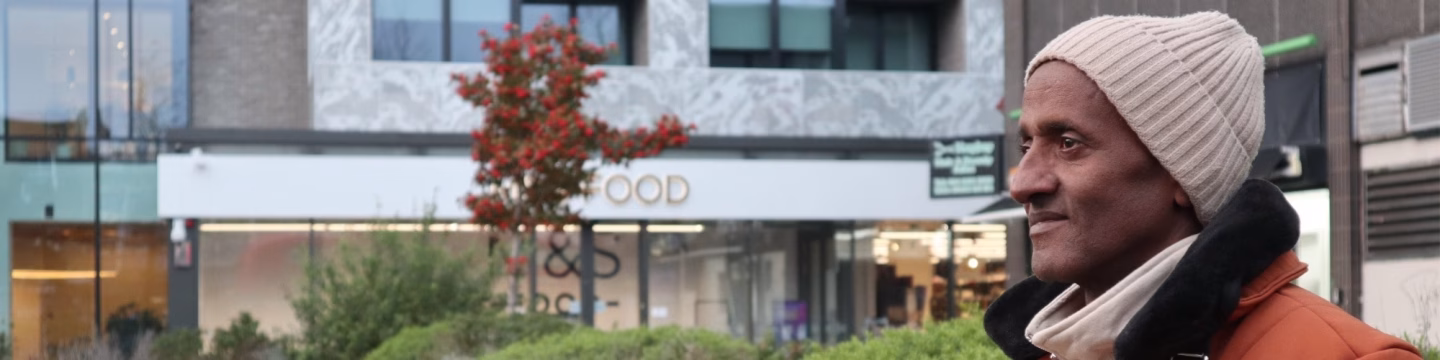 A man in winter clothes gazes into the distance, with autumn trees and buildings in the background.