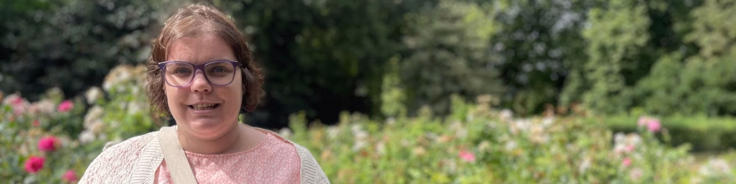 A woman is standing in a park, smiling, with flowers in the background.