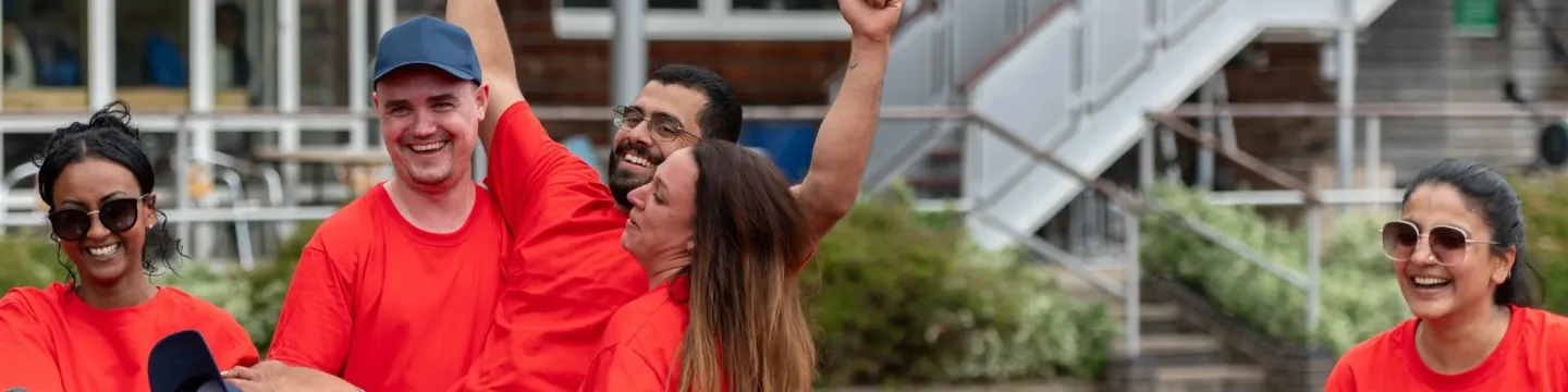A group of people smiles and cheers as they lift one group member in the air after winning a race on an athletics track.