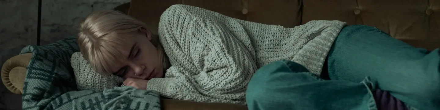 A young woman lies on a brown sofa, looking sad in a cold and lonely room with a white brick wall in the background.