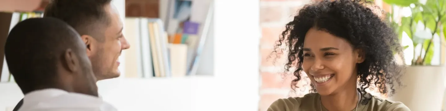 Three people are sitting around a table indoors, chatting and laughing.