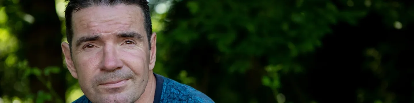 A man with a grin sits against a background of greenery.
