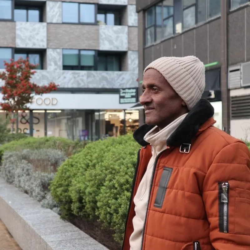A man in winter clothes gazes into the distance, with autumn trees and buildings in the background.