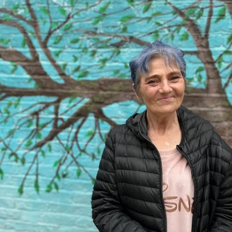 A woman is standing, smiling, with a colourful backdrop.