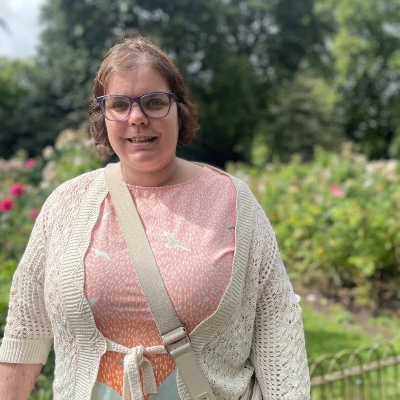 A woman is standing in a park, smiling, with flowers in the background.