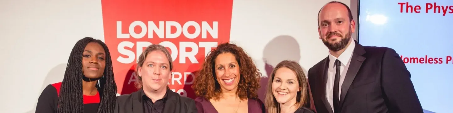 A group of four women and one man at an awards ceremony, smiling and celebrating after winning an award. They are dressed formally, standing on stage with a trophy in hand.