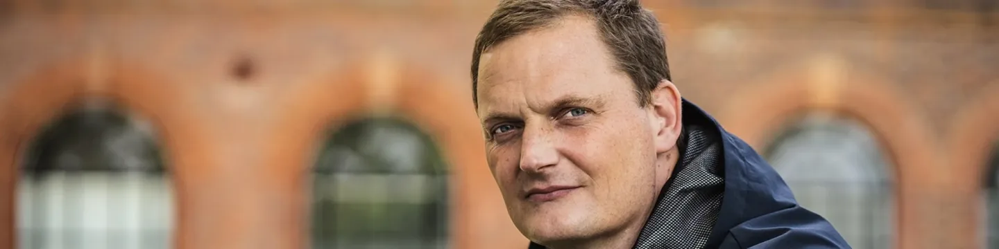 A man wearing a black jacket is seated in front of a building, appearing contemplative.