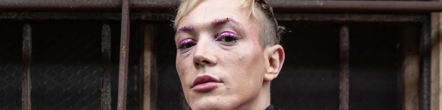 A man with blonde hair poses confidently with pink glitter on his face.