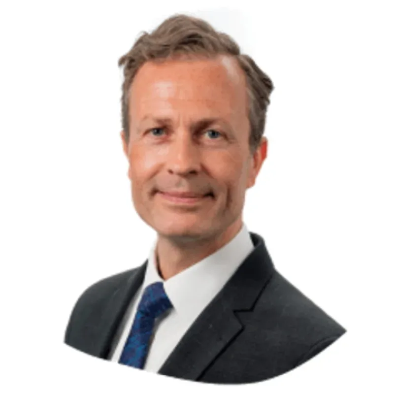 A man with short, slightly wavy hair wearing a black suit, white shirt, and a dark blue patterned tie.