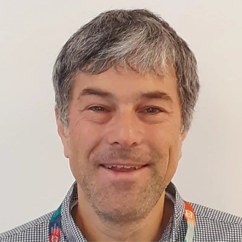 A man with grey hair, wearing a blue chequered shirt, stands confidently in front of a white wall.