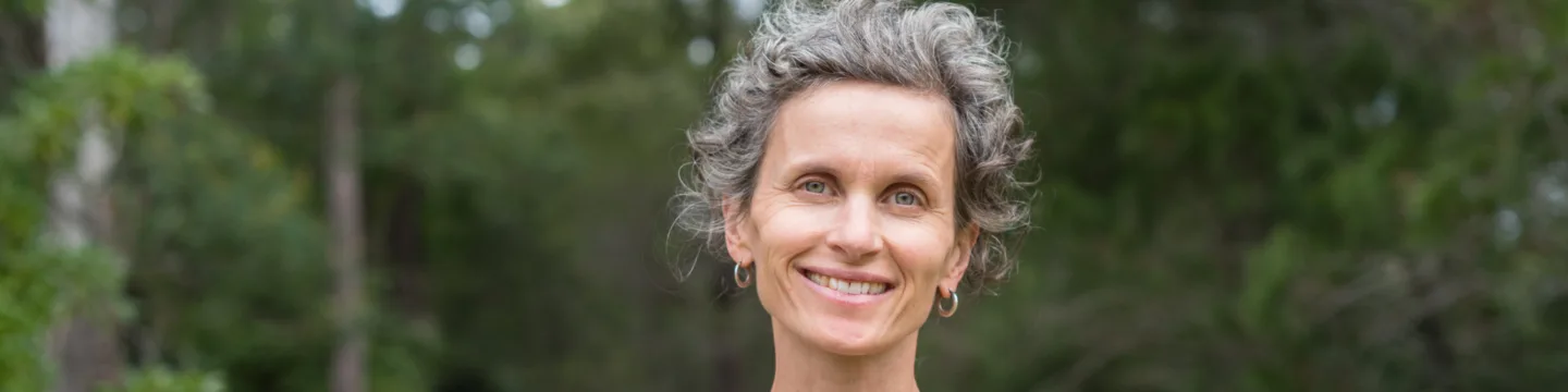A woman with grey hair and a gentle smile, wearing a blue shirt, stands in a forest.
