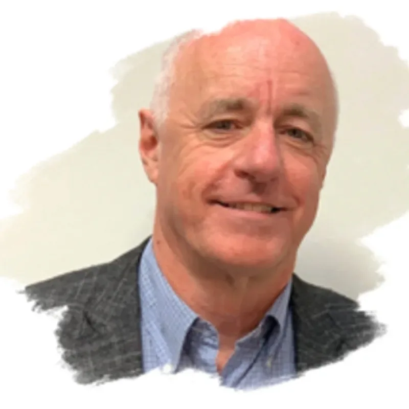 A senior man with short grey hair wearing a dark grey patterned suit and a light blue patterned shirt with the top button undone.