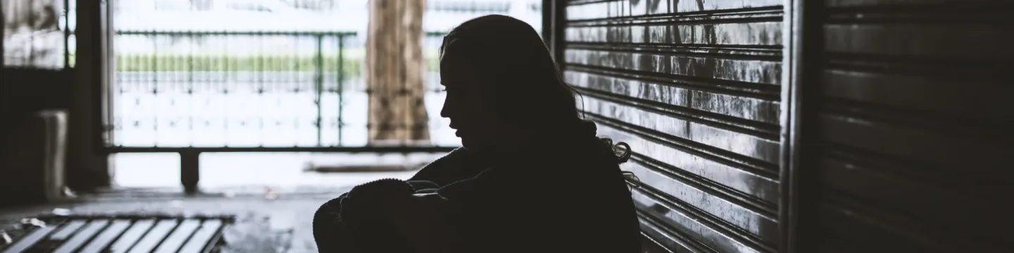 A silhouette of a woman sits on the floor in a dimly lit alleyway, her figure shadowed in solitude, capturing a moment of quiet vulnerability and reflection.
