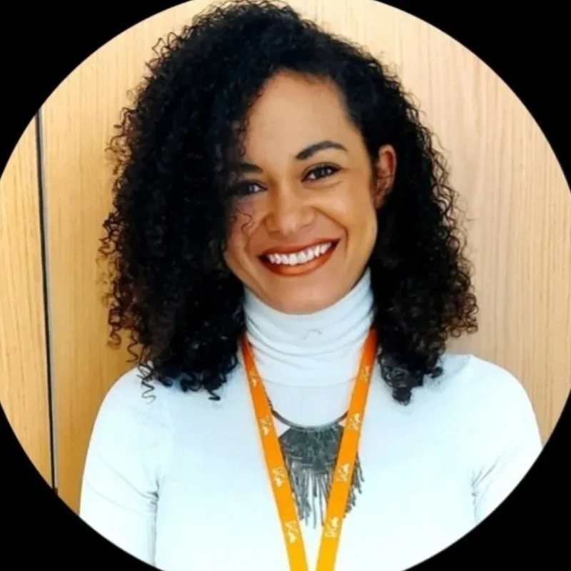 A woman with black curly hair, wearing a white turtleneck jumper, smiles.