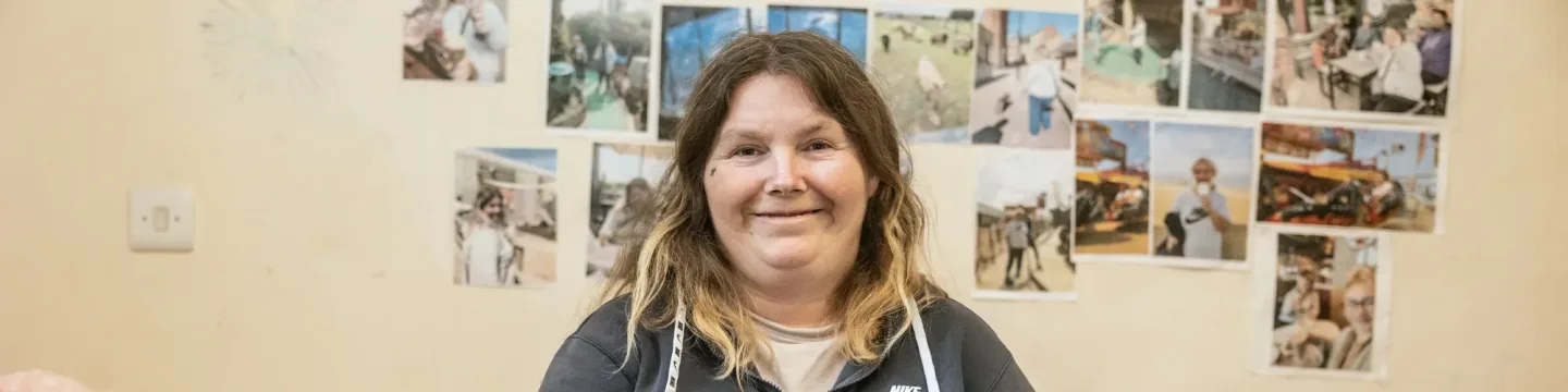 A woman sits on a bed surrounded by stuffed animals, with a cream wall covered in photographs, creating a cosy and homey environment.