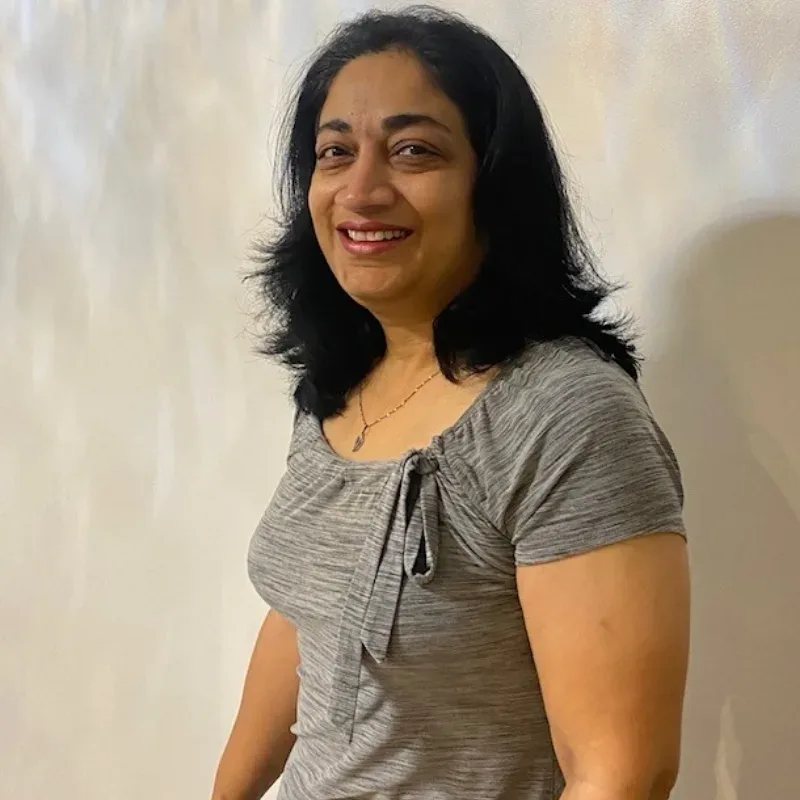 A woman with black hair wearing a grey shirt stands confidently before a plain white wall.