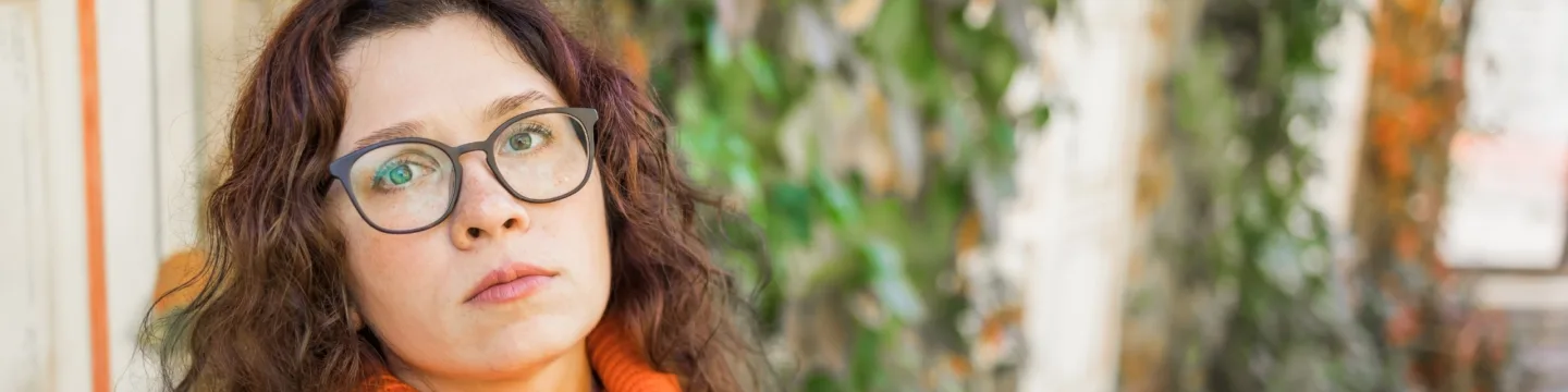 A woman with frizzy hair and glasses, dressed in an orange sweater, looks distant with a backdrop of greenery.