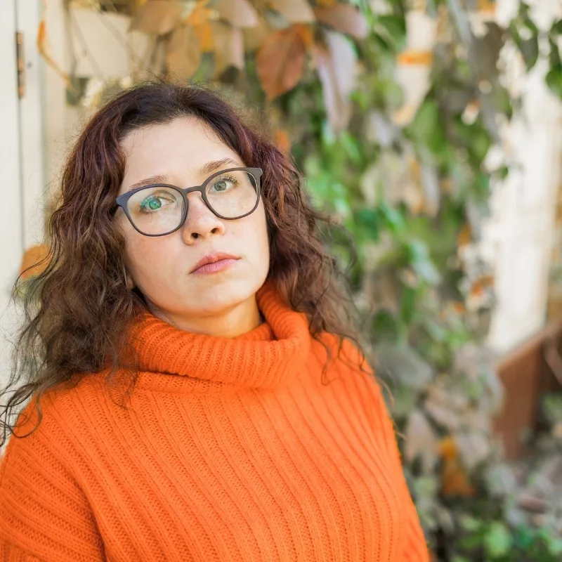 A woman with frizzy hair and glasses, dressed in an orange sweater, looks distant with a backdrop of greenery.