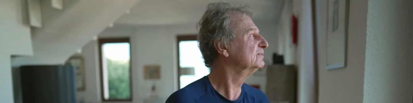 An elderly man gazes out the window, holding a cup of tea, his expression contemplative and emotionless.