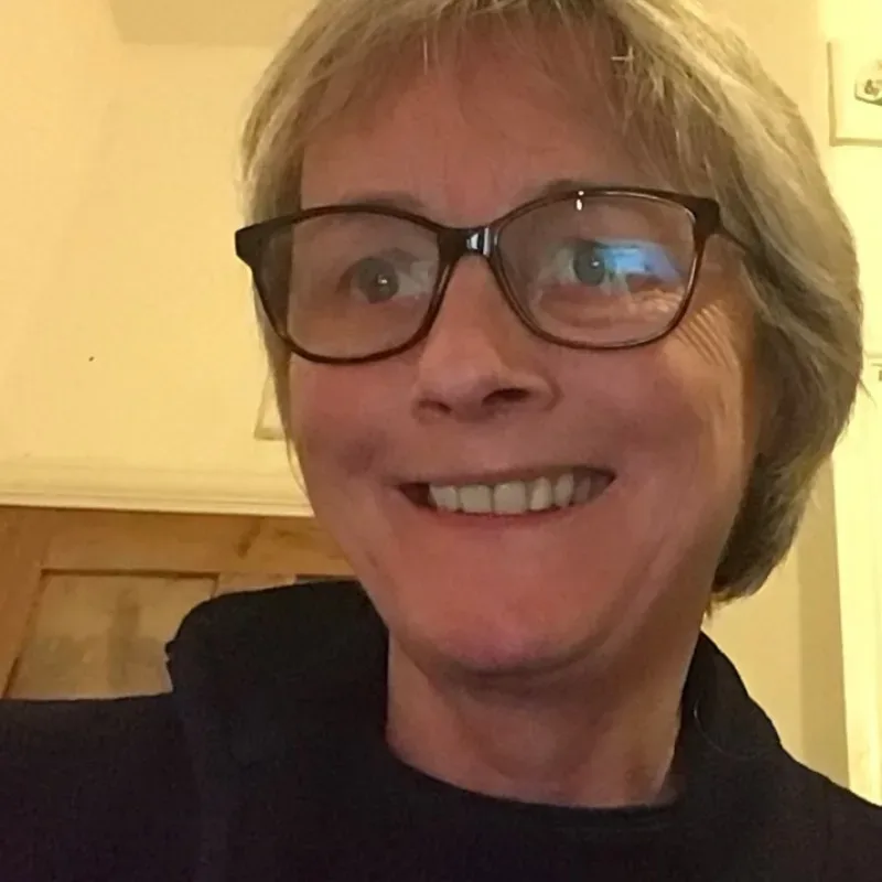 An older woman wearing glasses and a black shirt, smiling warmly against a cream-coloured wall background.