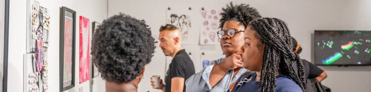 Three young women stand in an exhibition centre, facing and admiring an art piece on the wall. They are positioned close to one another, engaged and focused on the artwork in front of them, with the gallery's modern and well-lit interior visible in the background.