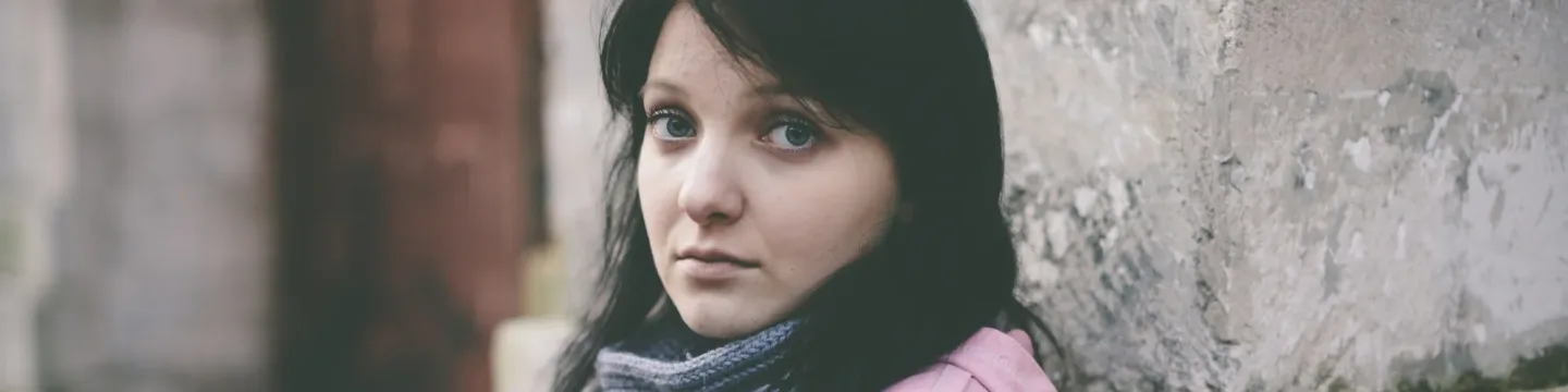 A woman dressed in winter clothing stands against a wall on the street, looking sad and lonely, with her head slightly bowed and arms crossed.