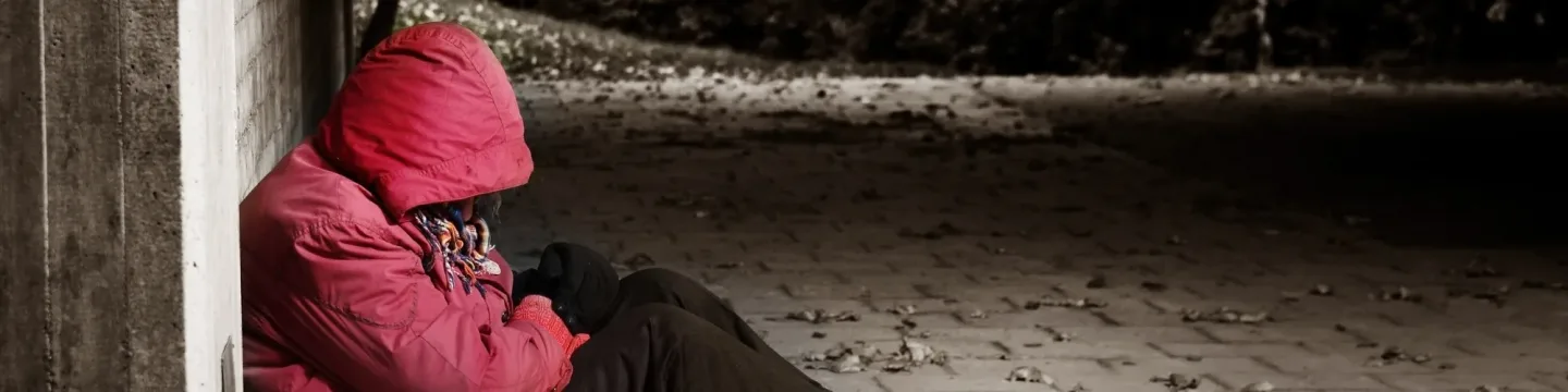 A homeless person wearing a red coat sits on the ground, reflecting a moment of hardship.