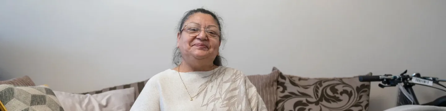 A smiling elderly woman with glasses, seated comfortably on a sofa with patterned cushions behind her. She is wearing a light-colored sweater and a delicate necklace.