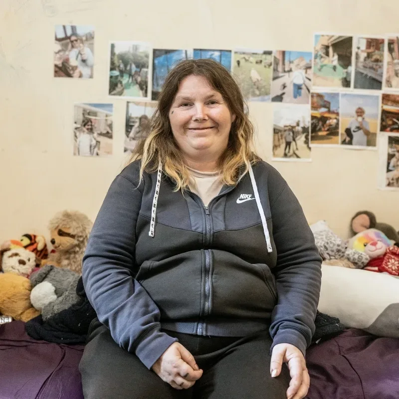 A woman sits on a bed surrounded by stuffed animals, with a cream wall covered in photographs, creating a cosy and homey environment.