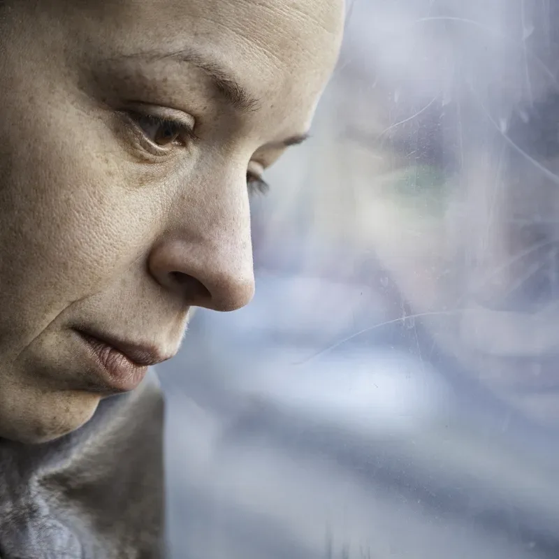 A woman sits quietly, staring at a frosted glass window, her expression thoughtful and distant.