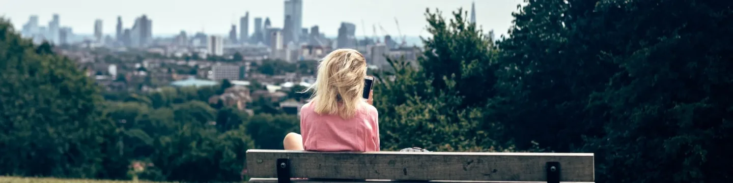 A woman with blonde hair sits on a bench in a park, looking at her phone with the London skyline in the distance.