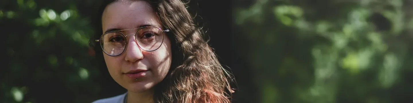 A young woman with brown wavy hair and large glasses stares the camera on a sunny day outside in a park