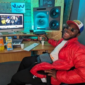 A young man smiles proudly at the camera whilst sitting at the computer producing music